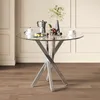 A round glass-top table with a chrome base, set against a paneled wall. Náhľad obrázku produktu