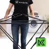 A person is assembling a black drying rack in a laundry room with a washing machine in the background. Podgląd zdjęcia produktu