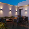 An outdoor patio at dusk, featuring modern furniture, plants, and wall-mounted lights. Produktbild-Vorschau