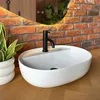 A white vessel sink with a black faucet is shown on a wooden countertop, with a brick wall and plants in the background. Náhled obrázku produktu