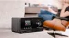 A black digital radio is placed on a sofa in a living room setting, with a person sitting in a chair in the background. Produktbild-Vorschau