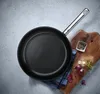 A black frying pan with a silver handle is placed on a gray surface next to a wooden cutting board with some ingredients. Podgląd zdjęcia produktu