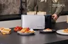 A person using a toaster in a kitchen setting with breakfast items. Voir les images du produit