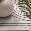 A close-up view of a textured beige pouf resting on a rug with a geometric pattern of concentric circles and horizontal lines. A shadow of a green leaf is cast on the rug. Produktbild-Vorschau