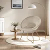 A beige velvet accent chair with gold metal legs is placed on a round white rug next to a small wooden side table with a vase of flowers. A floor lamp arches over the chair, and a framed picture hangs on the wall. Produktbild-Vorschau