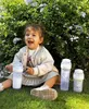 A happy toddler sitting on grass with baby bottles in front of a bush. Produktbild-Vorschau