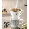 A pour-over coffee maker with a filter and ground coffee, with hot water being poured in, sits atop a striped mug. A measuring scoop and some golden foil packets are nearby, and a partially visible pastry is in the background. Produktbild-Vorschau