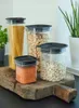 Four clear plastic storage containers with black lids are arranged on a wooden cutting board. The containers hold dry goods like spaghetti, beans, and lentils. A green plant is visible in the background. Anteprima dell’immagine prodotto