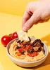 A hand is dipping a cracker into a bowl of hummus topped with sun-dried tomatoes and olives, with fresh tomatoes in the background. Produktbild-Vorschau