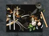 A black glass cutting board with a floral design, featuring a fork and knife on the left and a wooden spoon with spices and garlic on the right. Produktbild-Vorschau