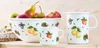 A white enamel bowl, mug, and pitcher decorated with fruit motifs, filled with fresh fruit, displayed on a wooden surface. Produktbild-Vorschau