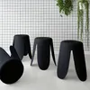 Four black stools are arranged on a light-colored floor in front of a white grid-patterned wall. Green plants hang from the top right of the image. Produktbild-Vorschau