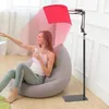 A woman is sitting on a beanbag chair, holding a mug, and reaching towards a laptop while a red light therapy panel shines on her. Produktbild-Vorschau