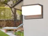 An outdoor wall lamp is mounted on a textured wall, with a garden scene in the background. Podgląd zdjęcia produktu