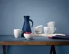 A navy blue carafe and white mugs and bowls are arranged on a blue surface, with raspberries in one bowl. Voir les images du produit
