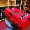 A red modular tool case with clamps attached, displayed on a stand in a retail environment with shelves of tool cases in the background. Anteprima dell’immagine prodotto
