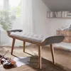 A tufted fabric bench with wooden legs is placed on a rug in a room with a window and bookshelves. Produktbild-Vorschau