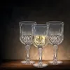 Three crystal wine glasses are arranged on a wooden surface against a dark, textured background. One glass contains a pale yellow liquid. Podgląd zdjęcia produktu
