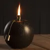 A black, spherical oil lamp with a lit wick and a gold pull chain sits on a wooden surface against a dark, textured background. Produktbild-Vorschau
