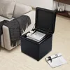 A black storage ottoman is open, revealing a book and a small stuffed animal inside. It is placed on a textured rug next to a sofa with a blanket draped over its arm. A magazine lies open on the rug in the foreground. Podgląd zdjęcia produktu