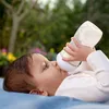 A baby lying on a blue blanket outdoors, drinking from a baby bottle. Anteprima dell’immagine prodotto