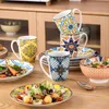 A table setting with plates, bowls, and mugs featuring a bohemian, hand-painted design. Náhled obrázku produktu