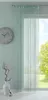 A mint-colored string curtain hangs in front of a window with a view of trees and sky. A clock is on the wall to the left, and a vase with branches and a decorative object are on the floor. Produktbild-Vorschau