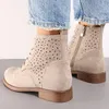 A close-up shot of a person wearing beige lace-up ankle boots with cut-out floral patterns, paired with blue denim jeans. Voir les images du produit
