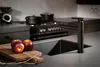 A modern kitchen scene with a black faucet, sink, and stove, featuring two red apples on a cutting board in the foreground. Produktbild-Vorschau