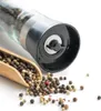 A close-up shot of a pepper grinder and a wooden scoop filled with peppercorns, set against a white background. Produktbild-Vorschau