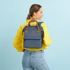 A woman wearing a yellow shirt and jeans is carrying a blue backpack. Produktbild-Vorschau