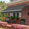 A gray awning extends over a brick wall with windows and flower boxes. A small table and chairs are set up on a brick patio in front of the awning, surrounded by pink flowers. Produktbild-Vorschau
