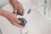 Hands washing a disassembled electric shaver under a running faucet in a sink. Náhled obrázku produktu