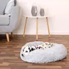 A Dalmatian dog is resting in a round, plush dog bed on a wooden floor, next to a grey sofa and a small white table with two decorative vases. Náhľad obrázku produktu