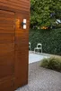 An outdoor wall light mounted on a wooden wall, with a garden setting in the background. Podgląd zdjęcia produktu
