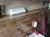 A white power strip with four outlets and two USB ports is placed on a shelf in a bathroom, next to a bottle of spray and a towel hanging on a hook. Produktbild-Vorschau