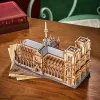 A wooden model of Notre Dame Cathedral sits on a wooden table next to a stack of papers and a red book. Náhled obrázku produktu