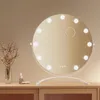A round makeup mirror with LED lights sits on a wooden dresser, next to a vase of dried flowers and some cosmetic products. Produktbild-Vorschau