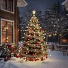 A decorated Christmas tree with lights and ornaments stands in the snow next to a wooden cabin, with more lights strung in the background trees. Podgląd zdjęcia produktu