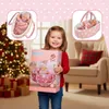 A young girl is holding a doll product box in front of a Christmas tree and a chair. There are inset images of the doll in its bassinet. Produktbild-Vorschau