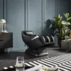 A black massage chair is shown in a modern living room setting with a large plant and a striped rug. Produktbild-Vorschau
