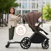 A young girl stands on a stroller board next to a stroller, outdoors on a paved walkway. Náhľad obrázku produktu