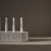 A white ceramic candle holder with fluted sides and four white taper candles is placed on a light gray surface. The background is a dark gray wall with soft shadows cast by the candles and holder. Produktbild-Vorschau