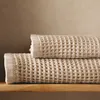 Two folded towels are stacked on a wooden shelf against a textured wall. Produktbild-Vorschau