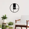 A modern black metal wall clock with a minimalist design is displayed on a white wall, with several potted plants and a chair in the foreground. Podgląd zdjęcia produktu