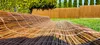 A rolled-up wicker mat lies on a green lawn, with a wooden fence and trees in the background. Podgląd zdjęcia produktu