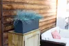 A rectangular planter with blue-green ornamental grass sits on a wooden cabinet next to a white outdoor sofa with a pink patterned cushion. Anteprima dell’immagine prodotto