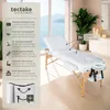 A white massage table set up in a bright, modern room with large windows and plants. Náhled obrázku produktu