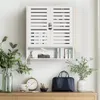 A white wall-mounted cabinet with slatted doors, displayed above a wooden dresser. The cabinet contains books and decorative items. The dresser holds a clock, a handbag, and plants. Podgląd zdjęcia produktu