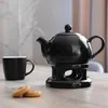 A black ceramic teapot with a lid and handle sits on a black stand with a tea light warmer. A black mug and two cookies are on the table next to it. Steam is rising from the teapot. Anteprima dell’immagine prodotto